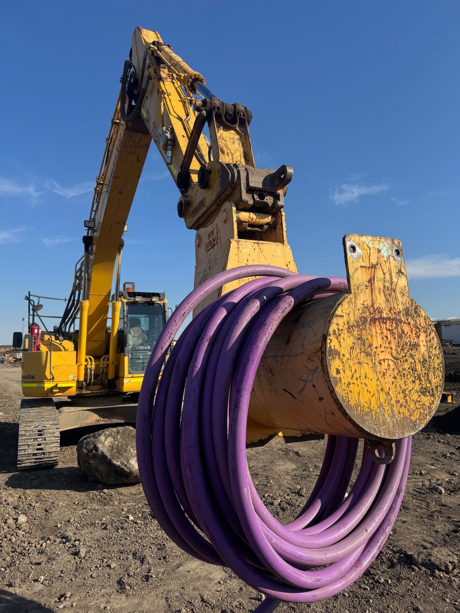 Yellow excavator with purple coiled tubing on a construction site under a blue sky.