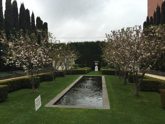A formal garden with a rectangular reflecting pool, lined with trees in bloom, hedges, and tall evergreens. Cloudy sky.