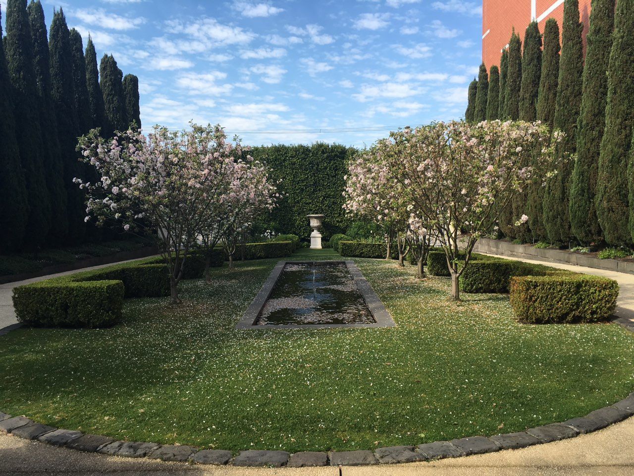 Formal garden with trees, a rectangular water feature, and trimmed hedges under a blue sky.