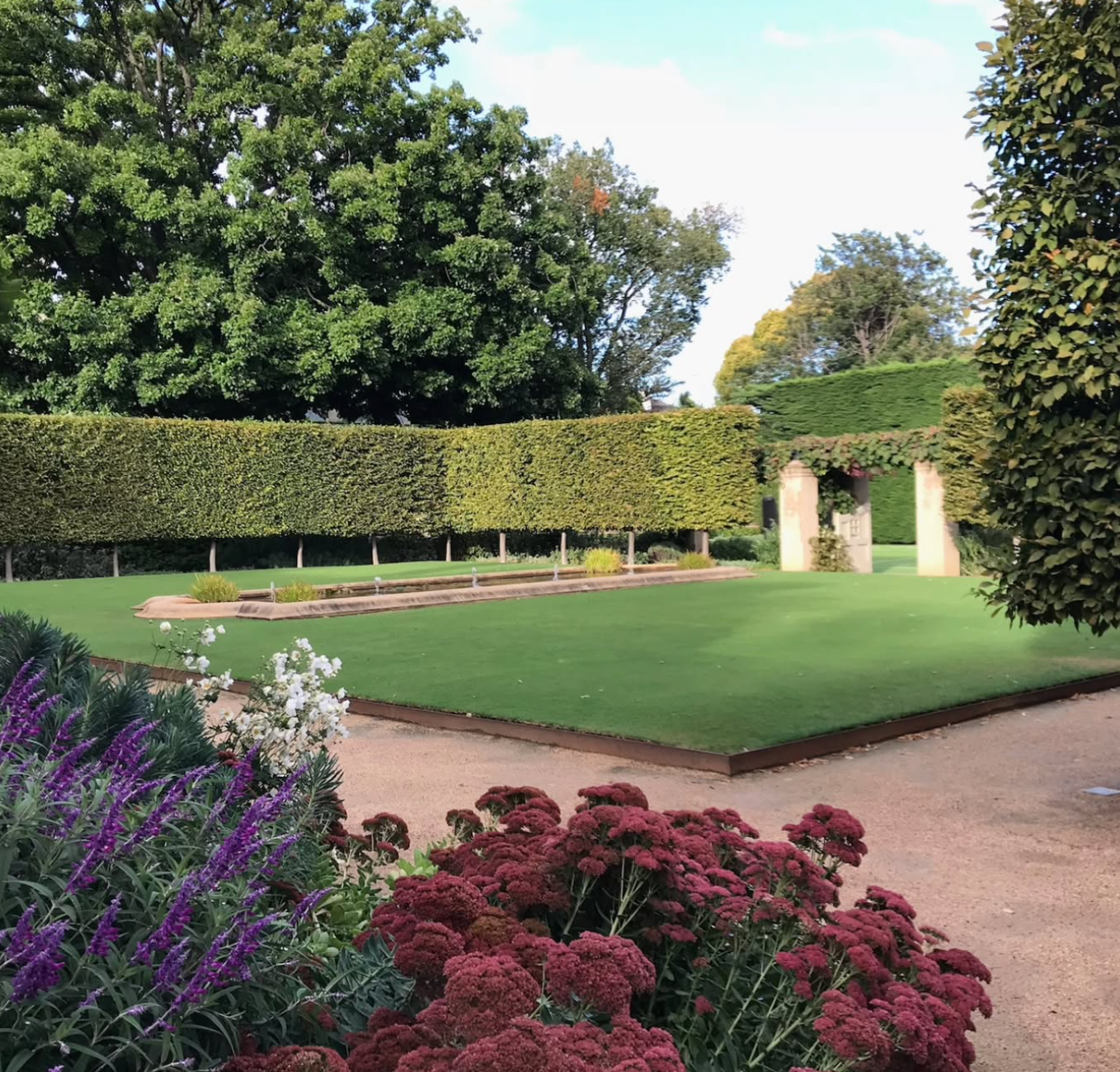 Lush garden with clipped hedges, green lawn, flower beds, and stone arches. Pathway leads to the garden on a sunny day.