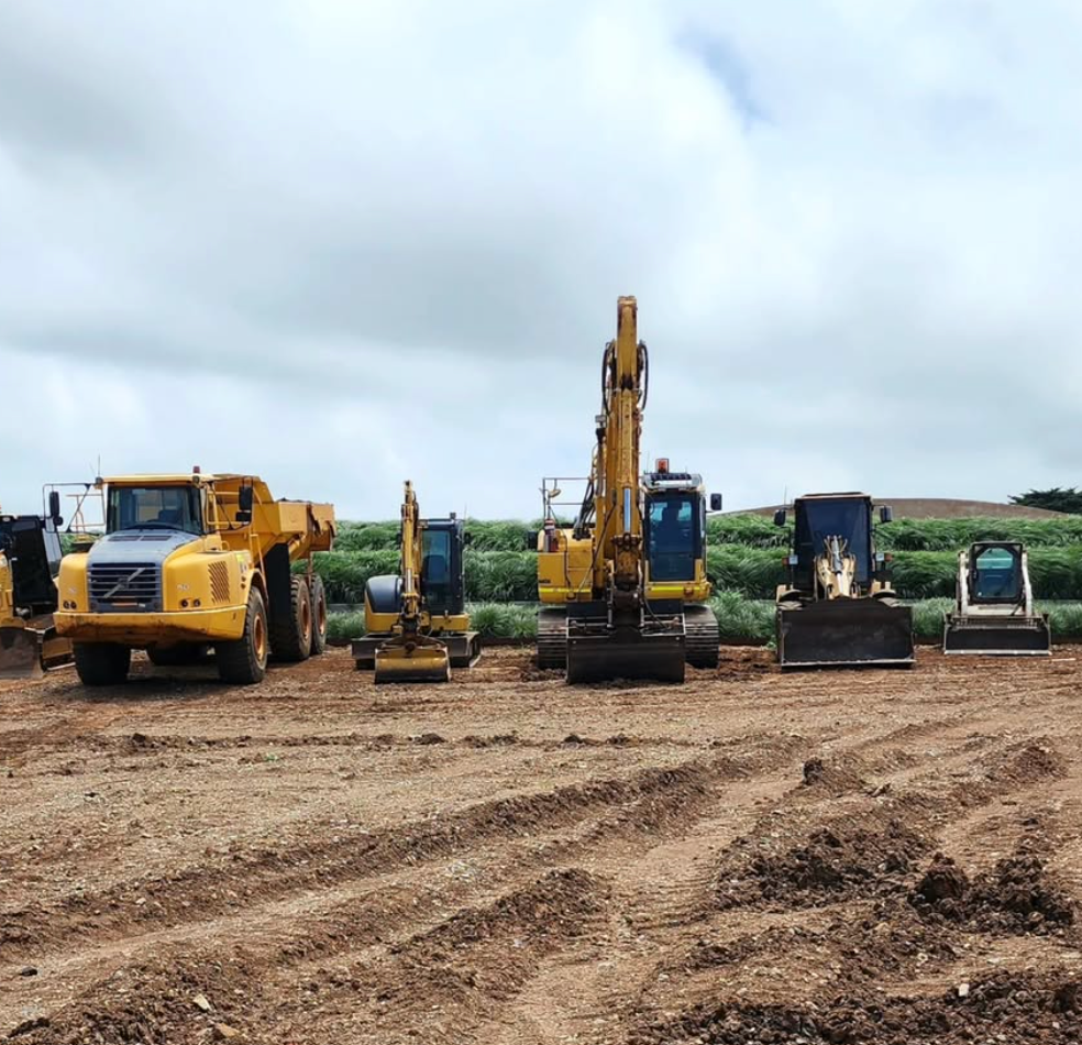 Construction equipment parked on a construction site: yellow dump truck, excavators, and a small tractor.
