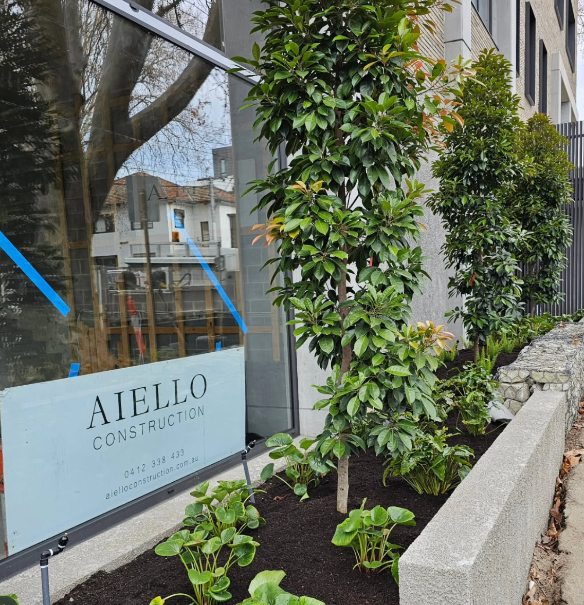 Green trees and bushes grow next to a modern building with a construction company sign.
