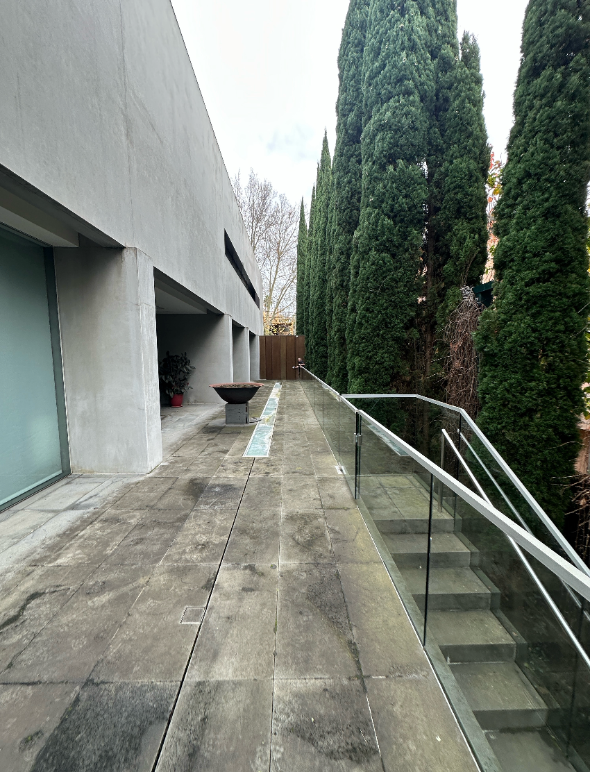 Narrow concrete walkway beside a building, with glass railing and tall evergreen trees on the right.