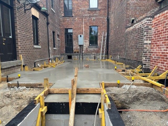 Concrete foundation under construction in a narrow brick alley, with wooden formwork and exposed rebar.