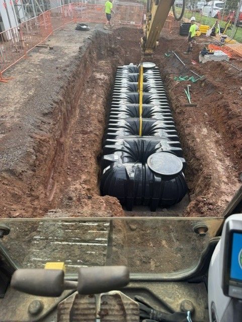 Construction trench with large black underground tank sections being installed, workers and machinery visible.