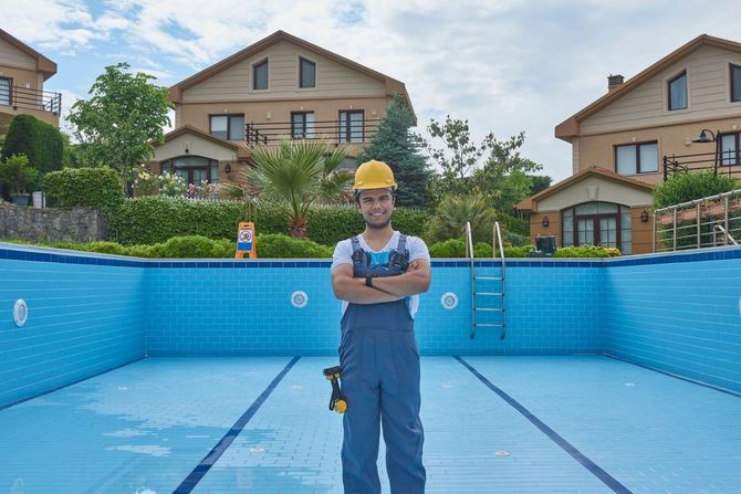 Worker standing in an empty blue swimming pool under construction, holding blueprints.
