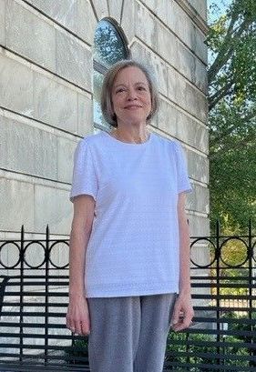 Deborah Crews wearing a white t-shirt and grey pants is standing in front of a building.