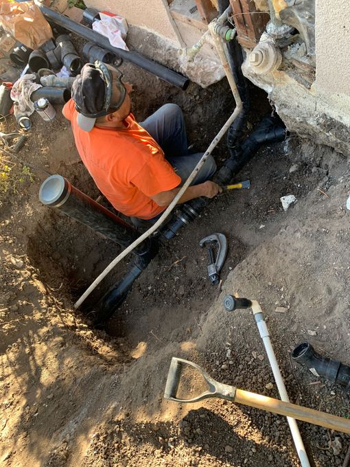 A worker in an orange shirt kneels in a trench, repairing a black plastic plumbing pipe next to a foundation.