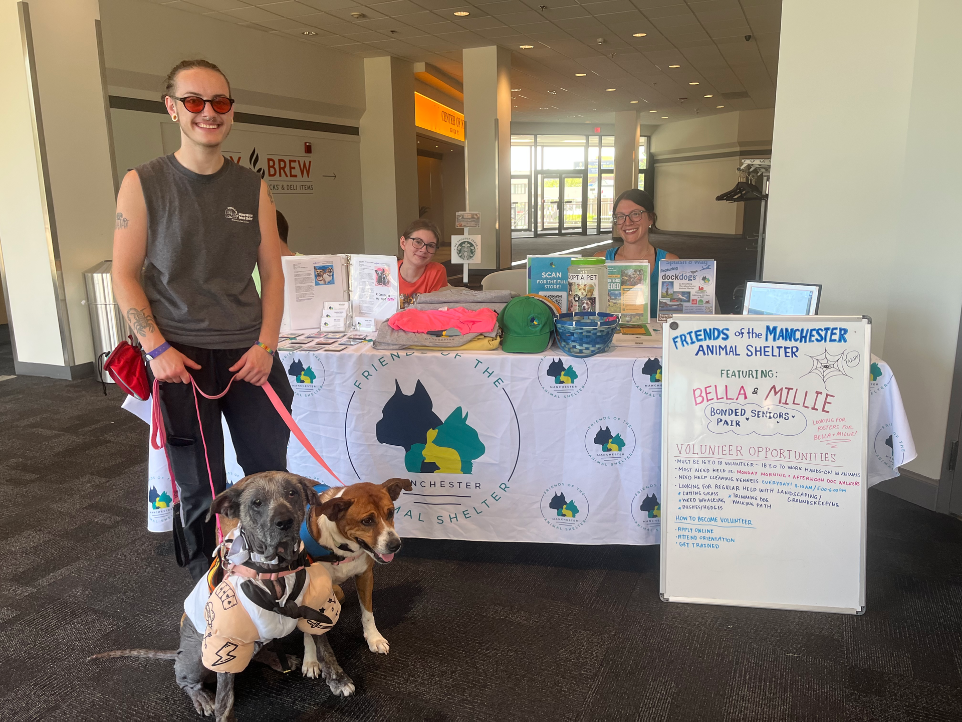 Two people stand behind a booth for an animal shelter, with two dogs sitting in the foreground.