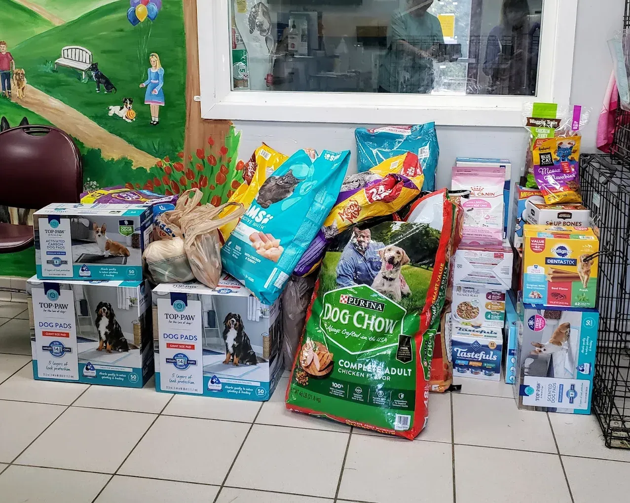 A collection of assorted dog food bags and boxes stacked indoors, set against a mural-painted wall.