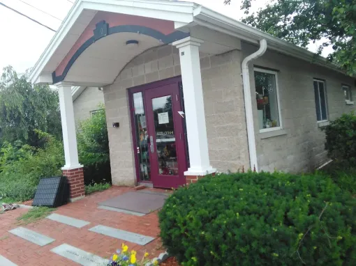 A small, light-colored building with a maroon door, a brick walkway, and a covered front porch supported by white pillars.