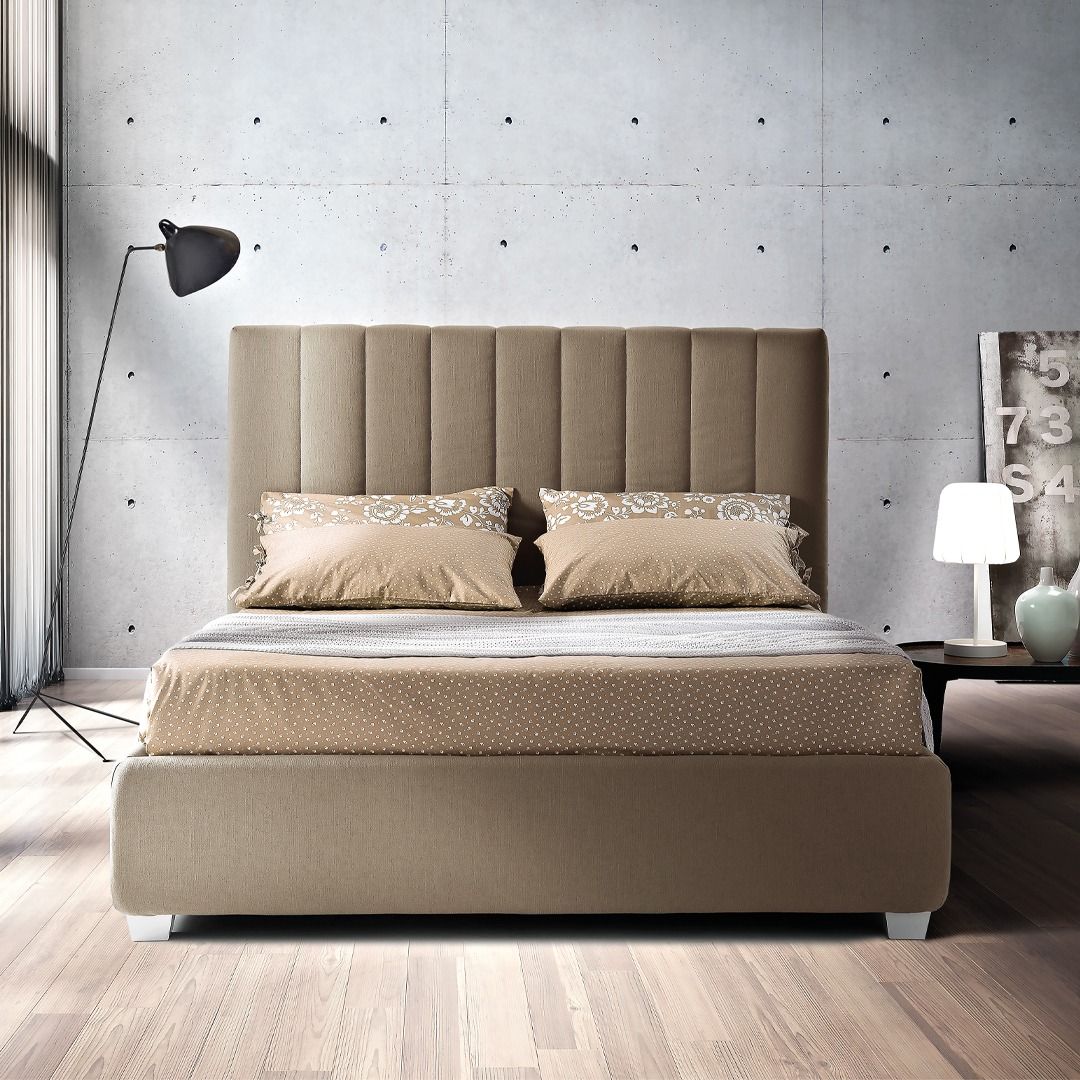 Un letto imbottito beige con una testiera alta a pannelli verticali, posizionato al centro di una stanza moderna con una parete in cemento.