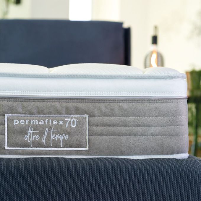 Primo piano di un materasso Permaflex 70 con pannello laterale grigio testurizzato, posizionato su una base blu scuro in una camera da letto illuminata da una luce soffusa.