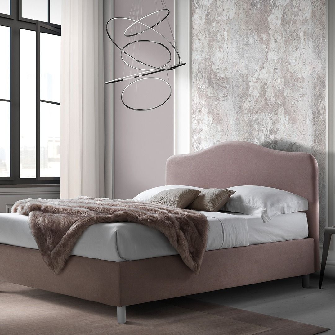 In una stanza, un letto imbottito color rosa antico con testiera curva, ricoperto da lenzuola bianche e un plaid in pelliccia sintetica marrone.