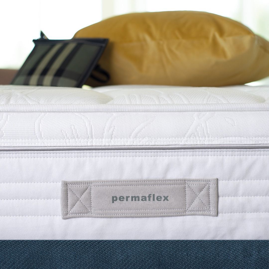 Primo piano del bordo di un materasso Permaflex bianco con maniglia in tessuto, con un cuscino giallo e una tasca nera sul retro.