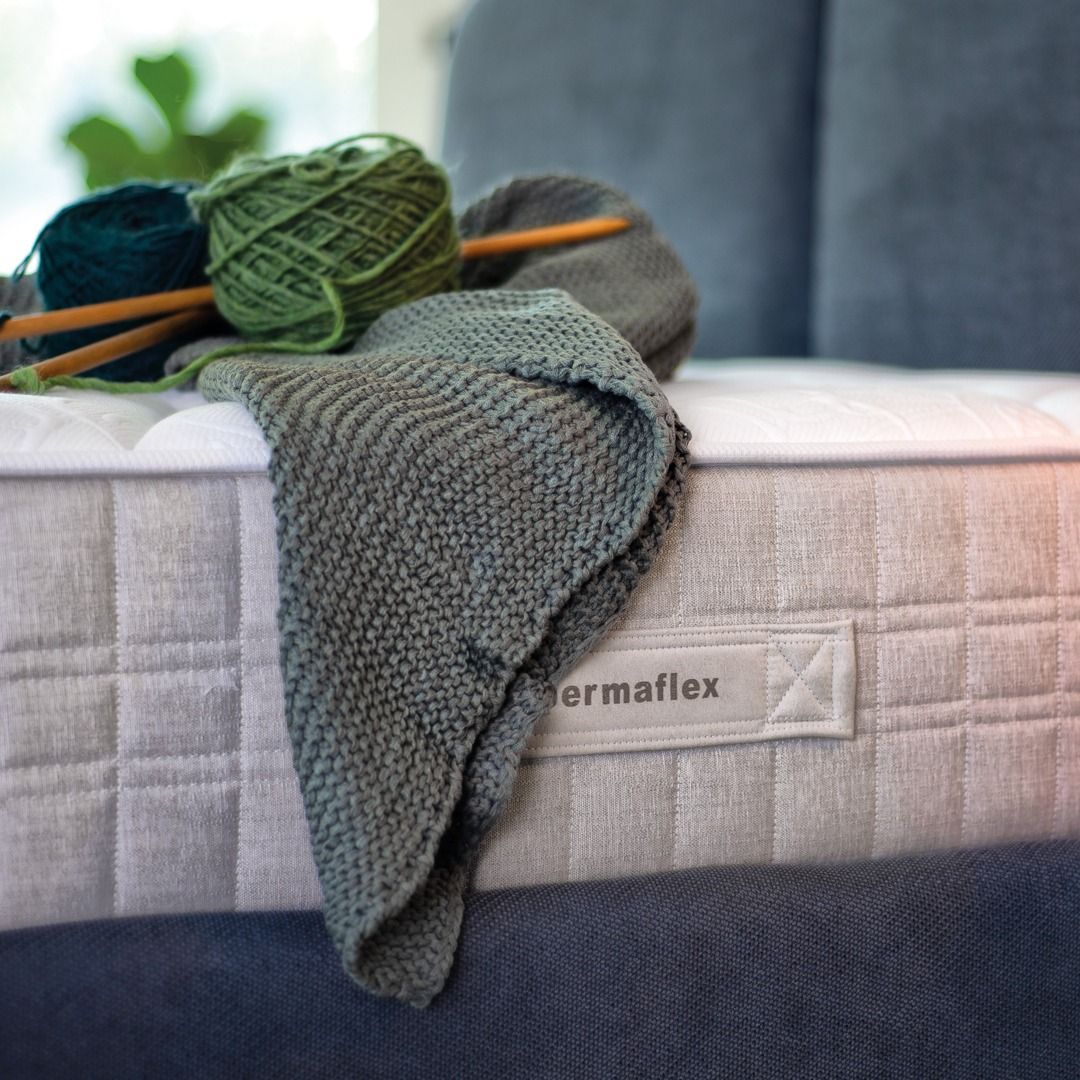 Una morbida coperta lavorata a maglia color verde salvia e gomitoli di lana con ferri di legno poggiano su un materasso grigio chiaro dalla texture particolare.