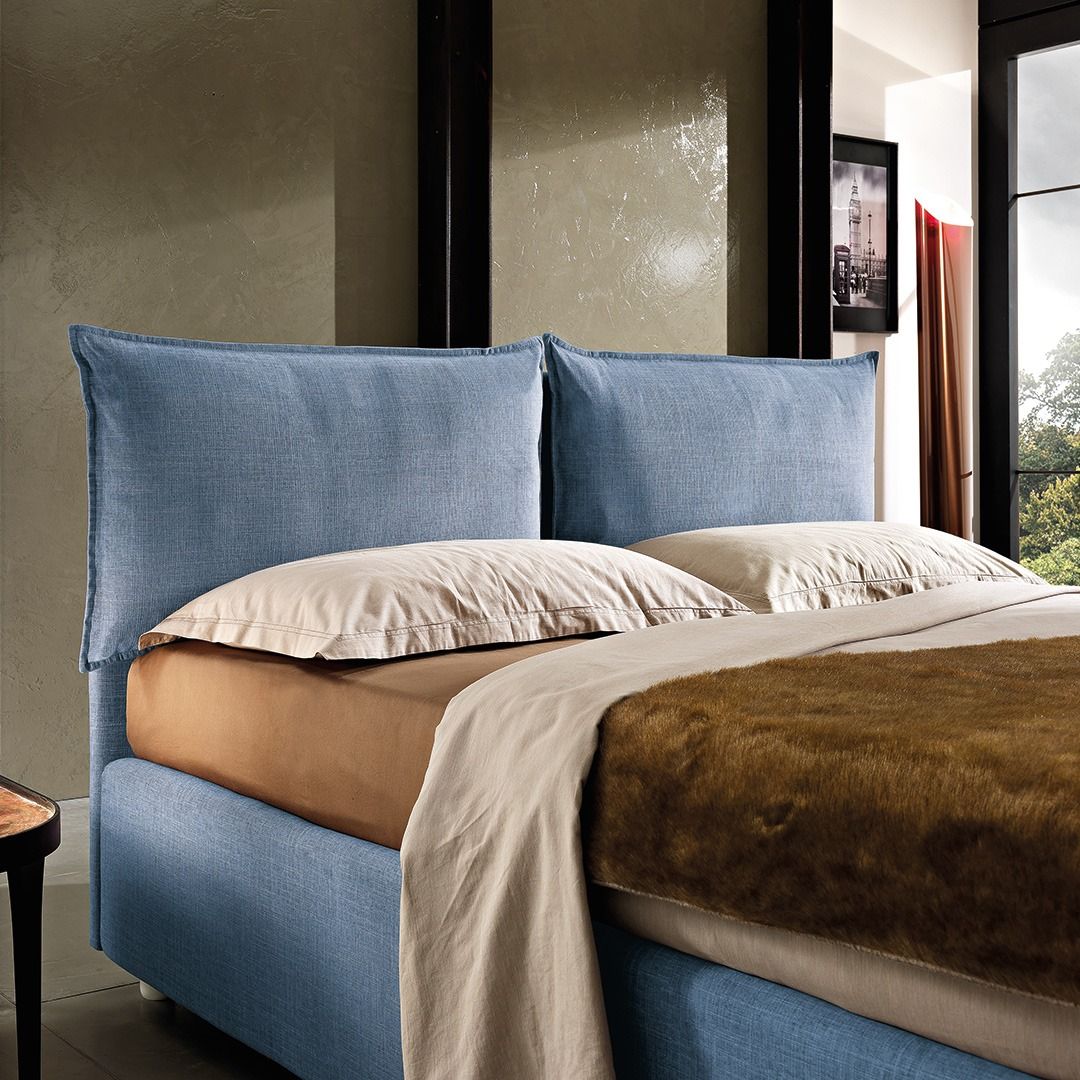 Una struttura letto imbottita di colore blu con due grandi cuscini rettangolari per la testiera, cuscini beige e una coperta marrone.