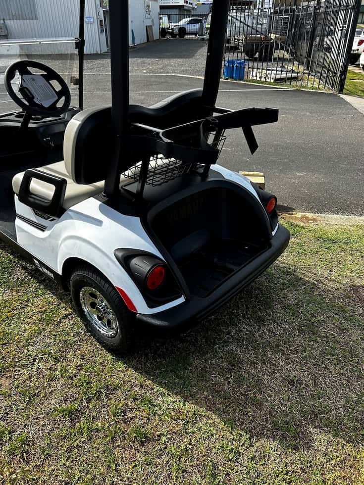 A White Golf Cart With a Rear-facing Seat on a Grassy Area Near a Paved Lot
