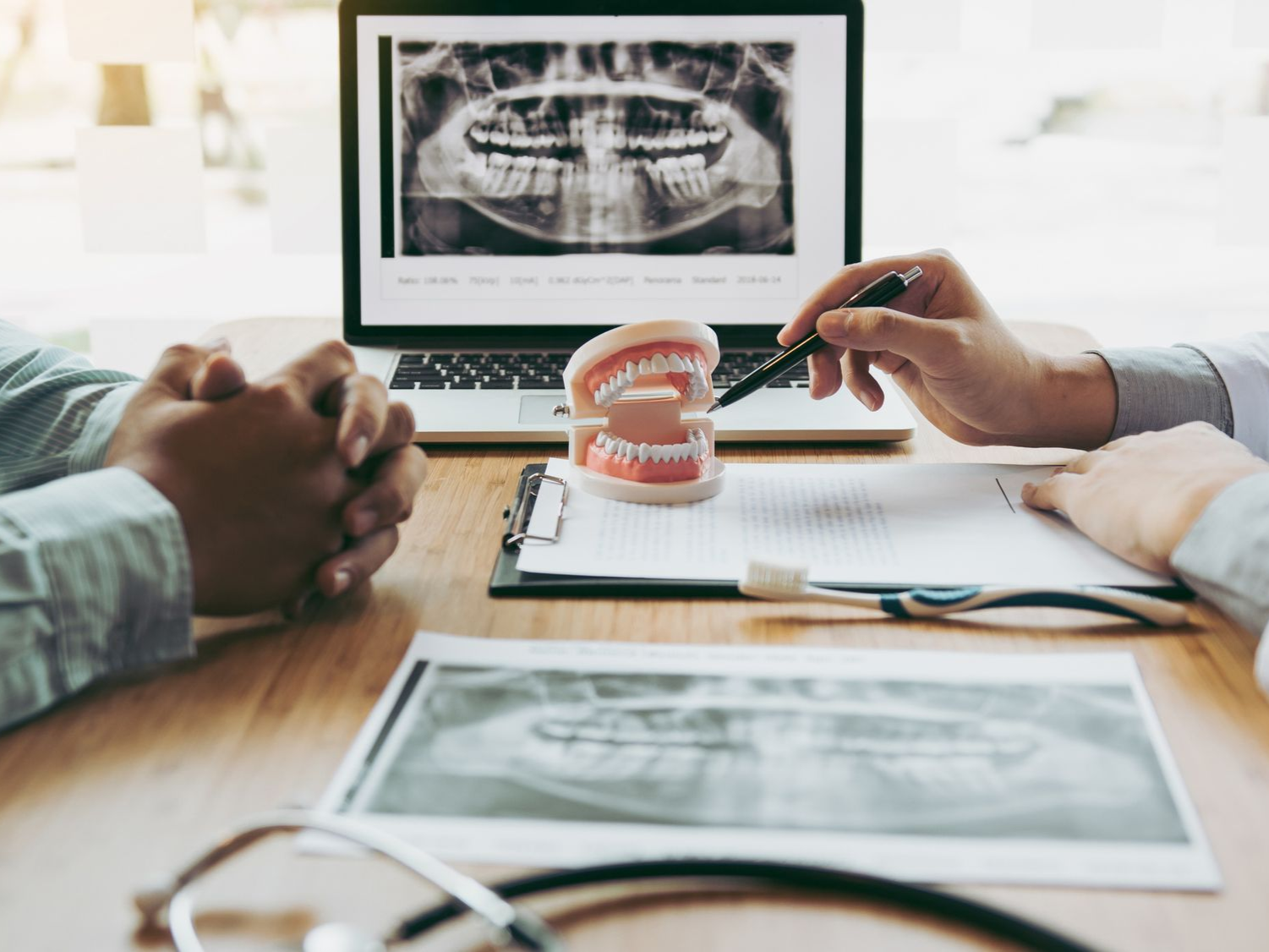 Dentist showing patient dental X-ray & model teeth, explaining treatment plan. Office setting, wooden desk.