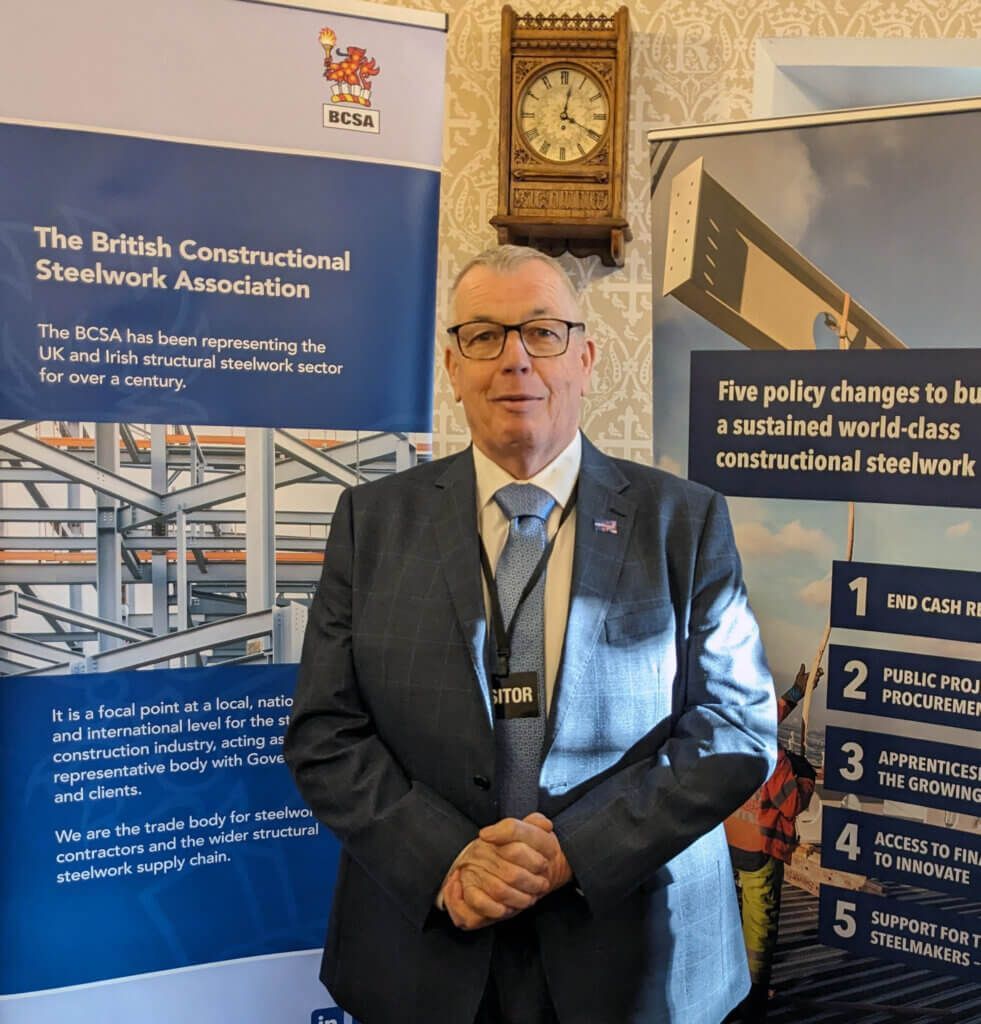 A man in a suit and tie is standing in front of a sign that says the british constructional steelwork association