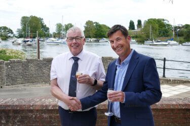 Two men are shaking hands in front of a body of water.