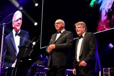 Two men in tuxedos are standing on a stage in front of a large screen.