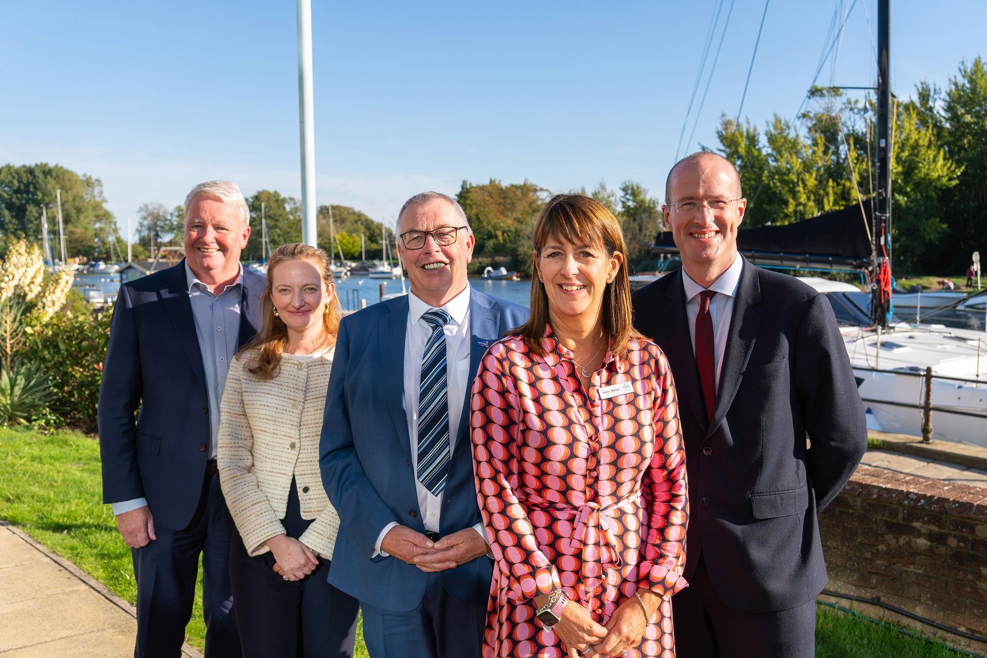 Five people posing outdoors near a harbor. They wear business attire and smile at the camera. Sunny day, boats in the background.