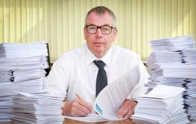 A man is sitting at a desk surrounded by stacks of papers.