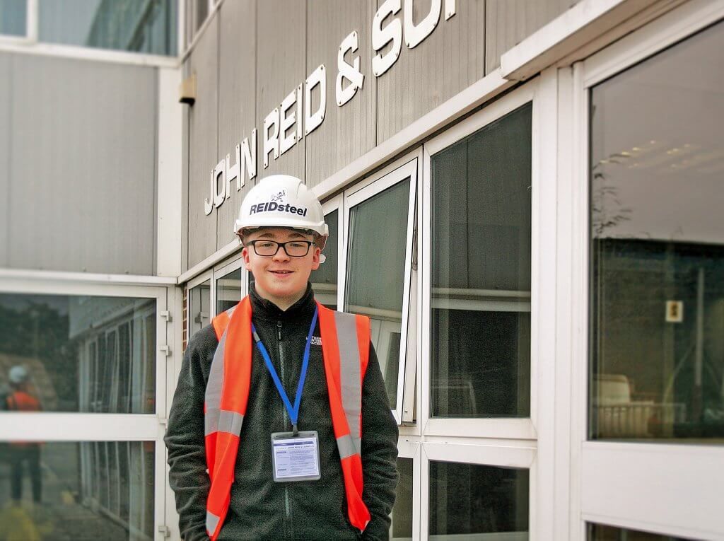 A man wearing a hard hat and safety vest stands in front of a building that says light red & sun