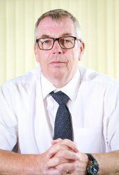 A man wearing glasses and a tie is sitting at a table with his hands folded.