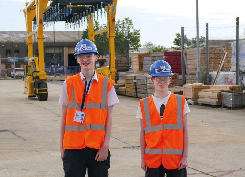 Two boys wearing hard hats and orange vests are standing in front of a yellow crane.