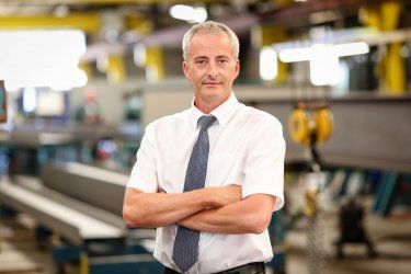 A man in a white shirt and tie is standing in a factory with his arms crossed.