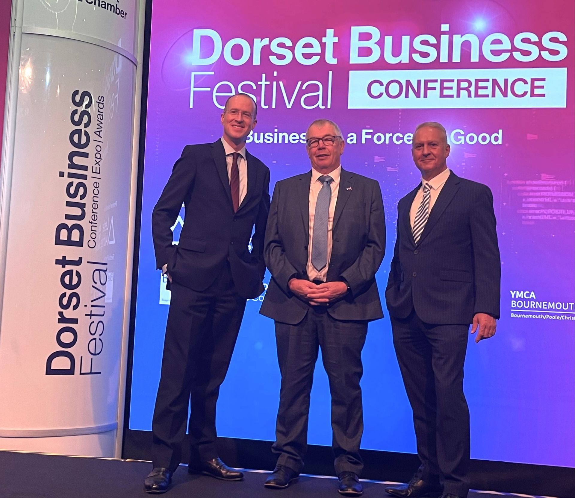 Three men are standing in front of a screen that says dorset business festival conference