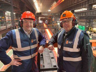 Two men wearing hard hats and safety vests are standing next to each other in a factory.