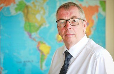 A man wearing glasses and a tie is standing in front of a map of the world.