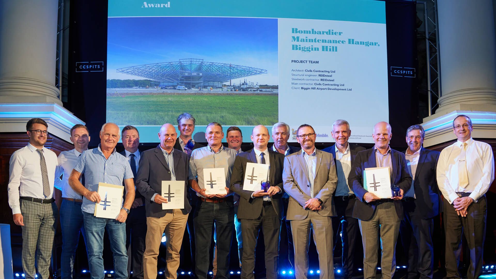 A group of men are standing in front of a large screen holding awards.