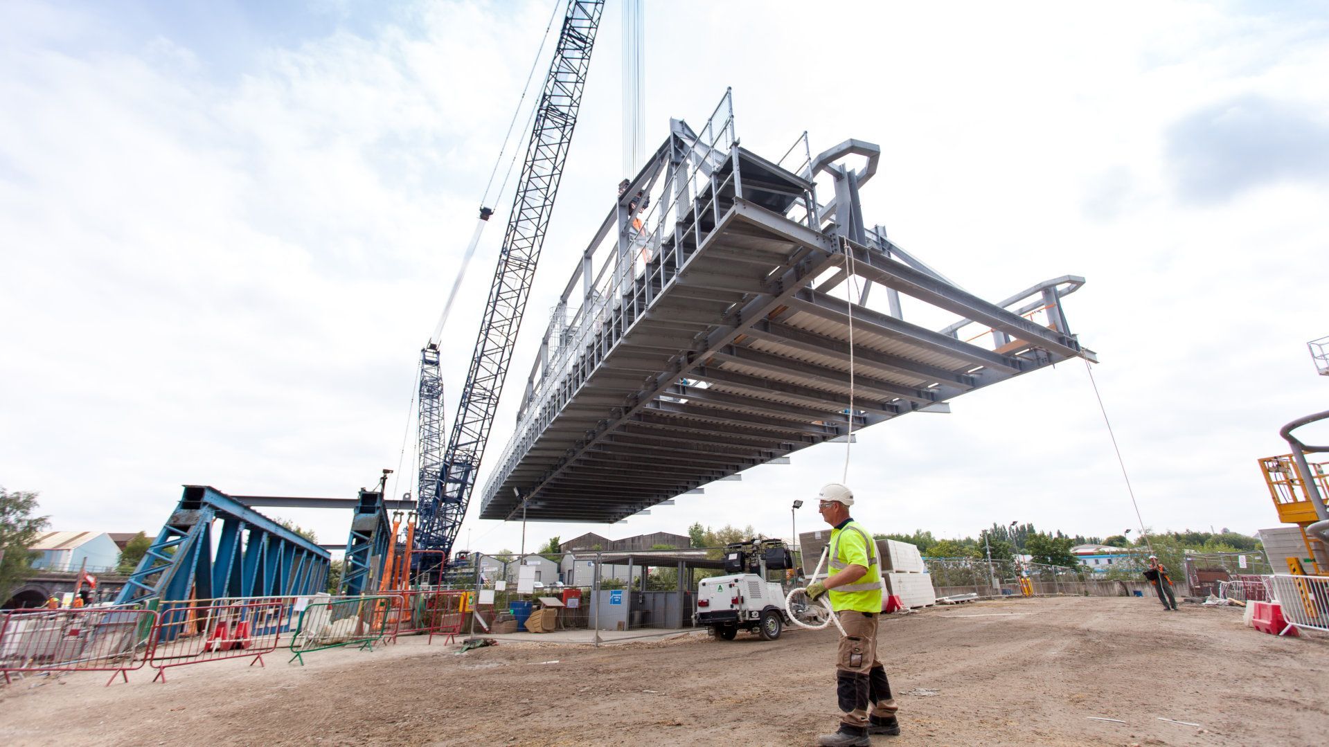 A man is standing in front of a large metal structure being lifted by a crane.