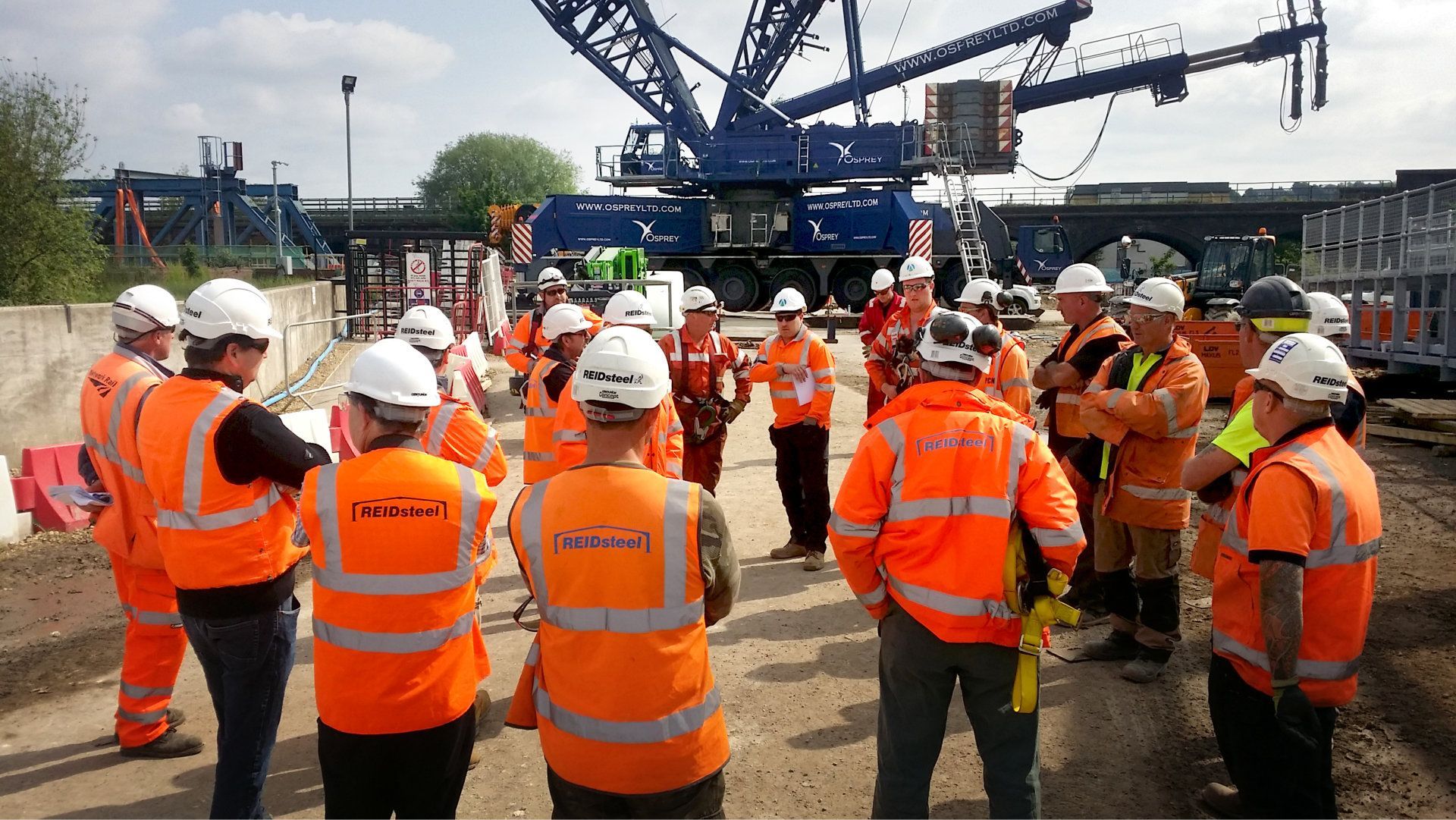 A group of construction workers wearing orange vests and hard hats are standing in a circle.