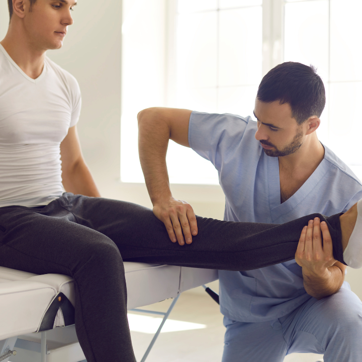 Physiotherapist examines a patient's leg on a white medical table in a sunny room.