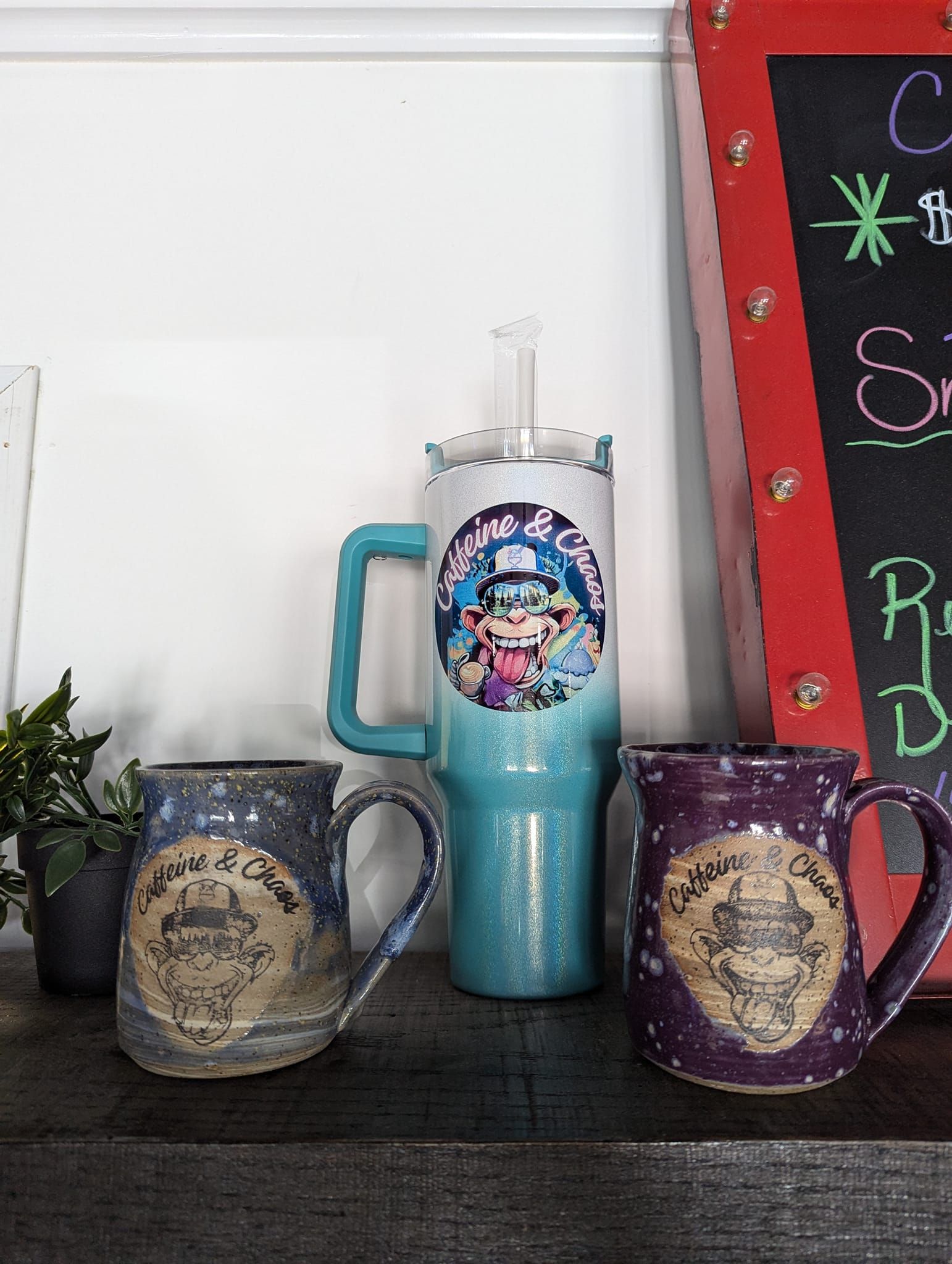Three mugs on a shelf: two blue and purple mugs with a label and a tall teal tumbler with a straw.