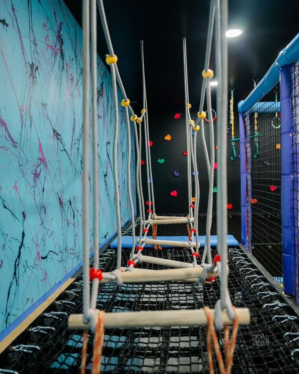 Rope bridge structure in an indoor playground, with wooden rungs, netting below, and colorful walls.