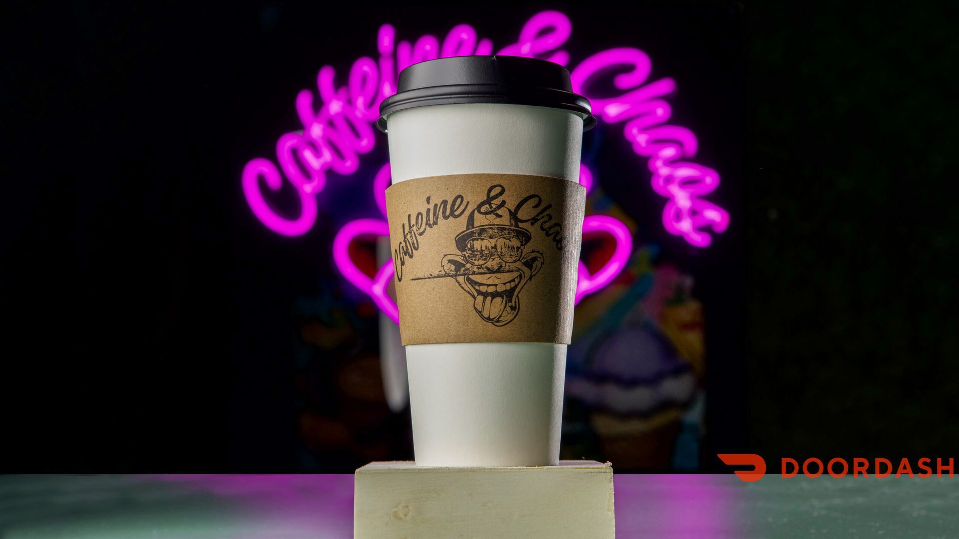 Coffee cup with branded sleeve in front of neon “Caffeine & Chaos” sign, DoorDash logo at bottom.