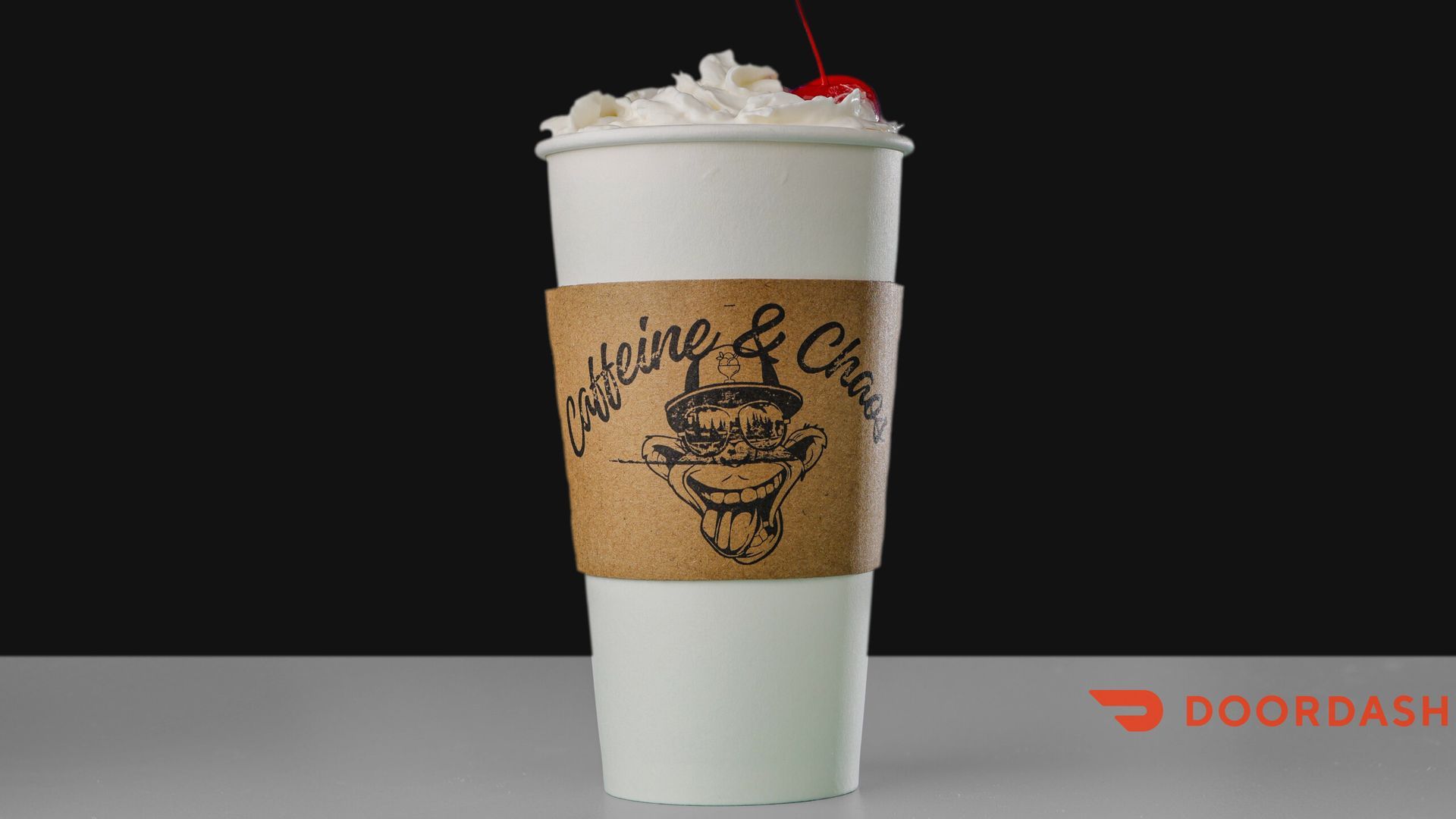 White coffee cup with whipped cream and cherry, brown sleeve with logo, on a white surface.