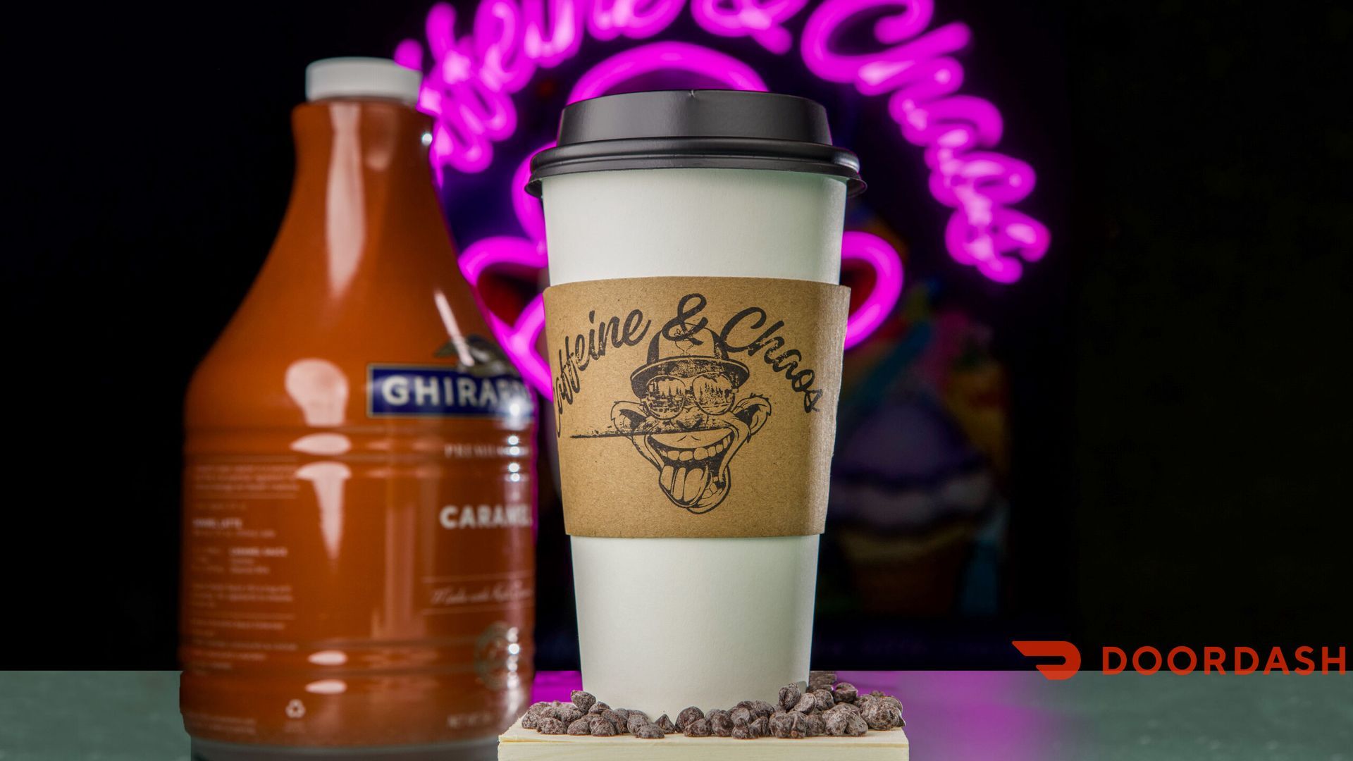Coffee cup with caramel syrup bottle and neon sign in background.