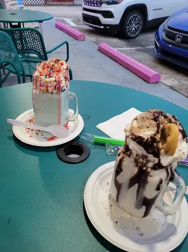 Two elaborate milkshakes on a green table, outdoors. One with sprinkles, the other with chocolate and a cookie.