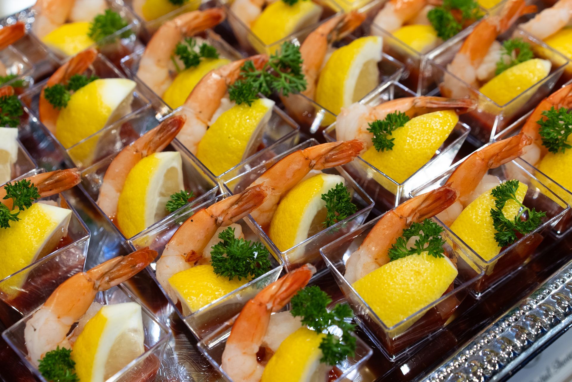 Shrimp cocktail appetizers in clear square cups with lemon slices and parsley garnish on a silver platter.
