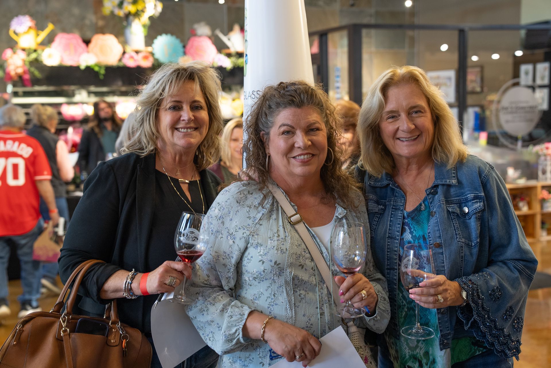 Three women smiling and holding wine glasses in a shop, one by a white column.