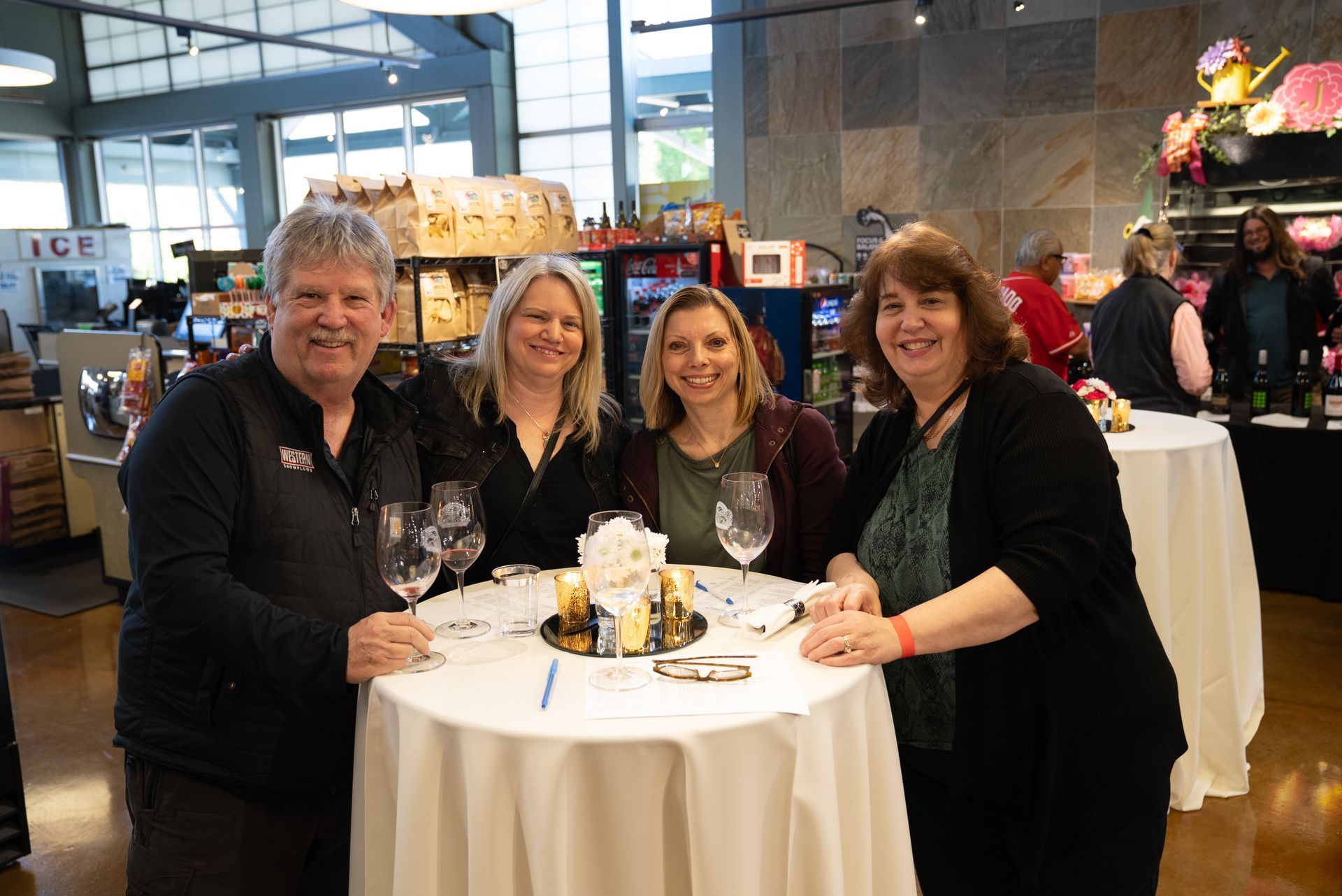 Four people smile around a small table with wine glasses. Indoor setting with food and drinks visible.