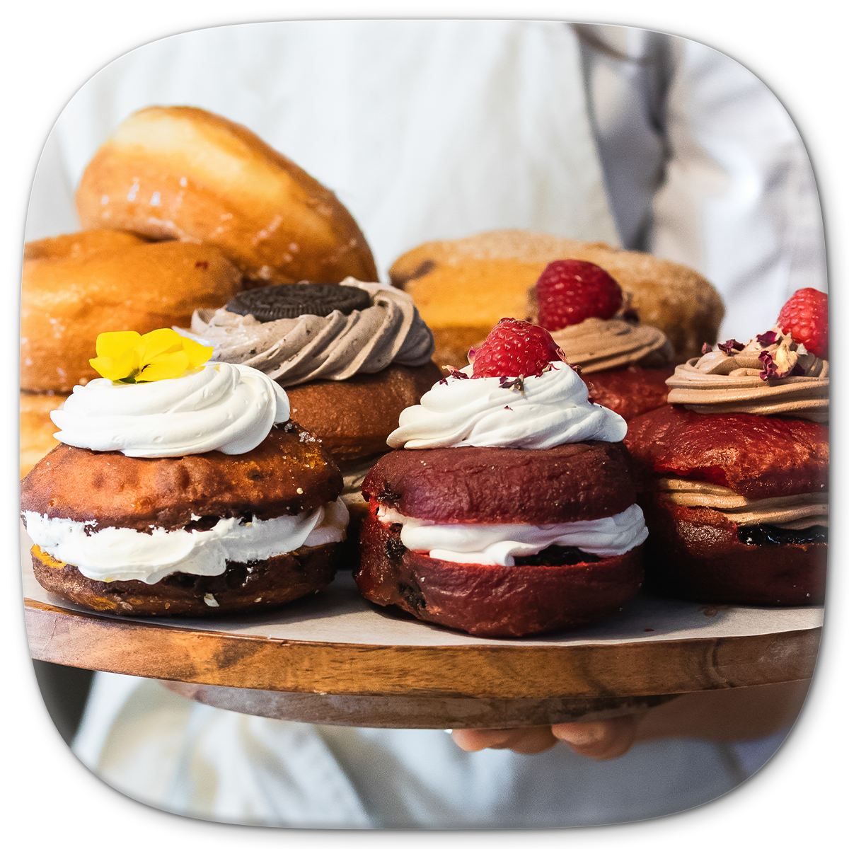 A selection of decorated donuts on a wooden board, held by a person in a white apron.
