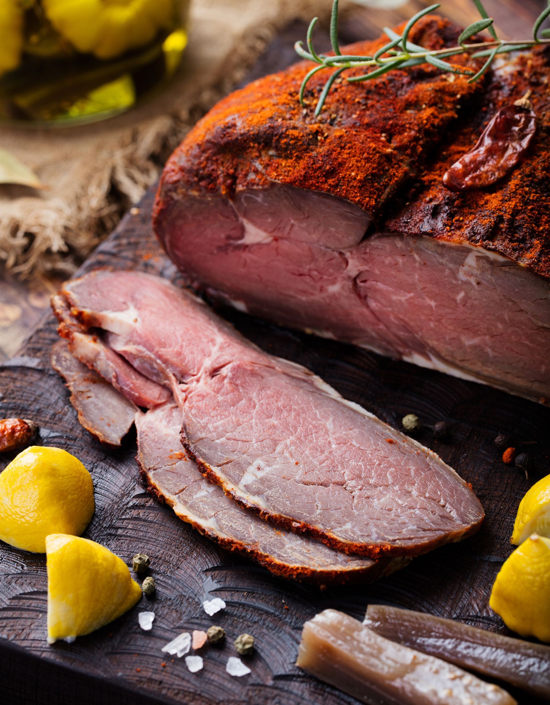 Sliced cured meat on a wooden board, seasoned with paprika, lemons, and herbs.